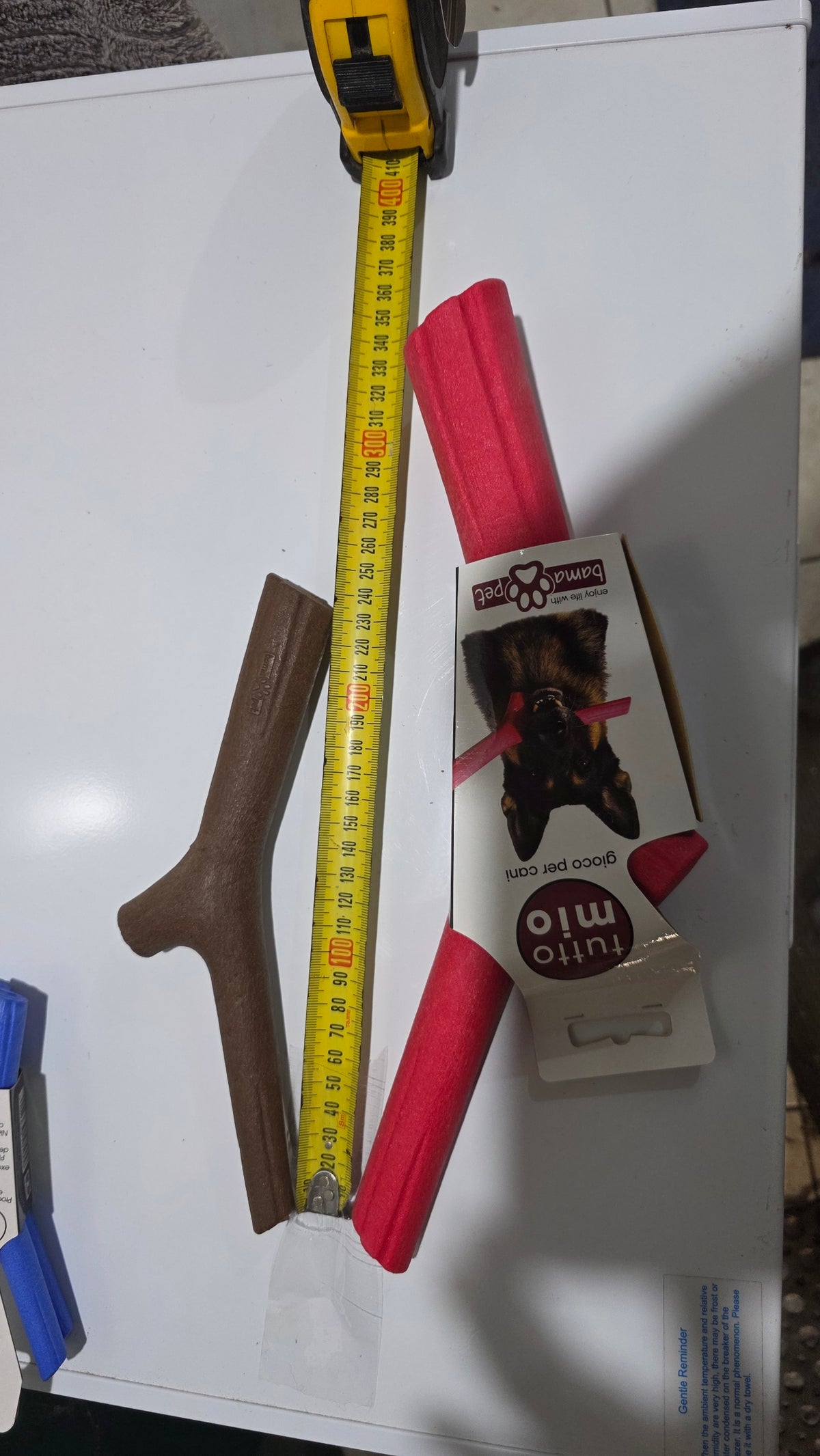 Packaged and unpackaged red, brown, and blue Bama floating stick dog toys arranged on a white surface next to a yellow tape measure demonstrating size differences across small, medium, and large variants.