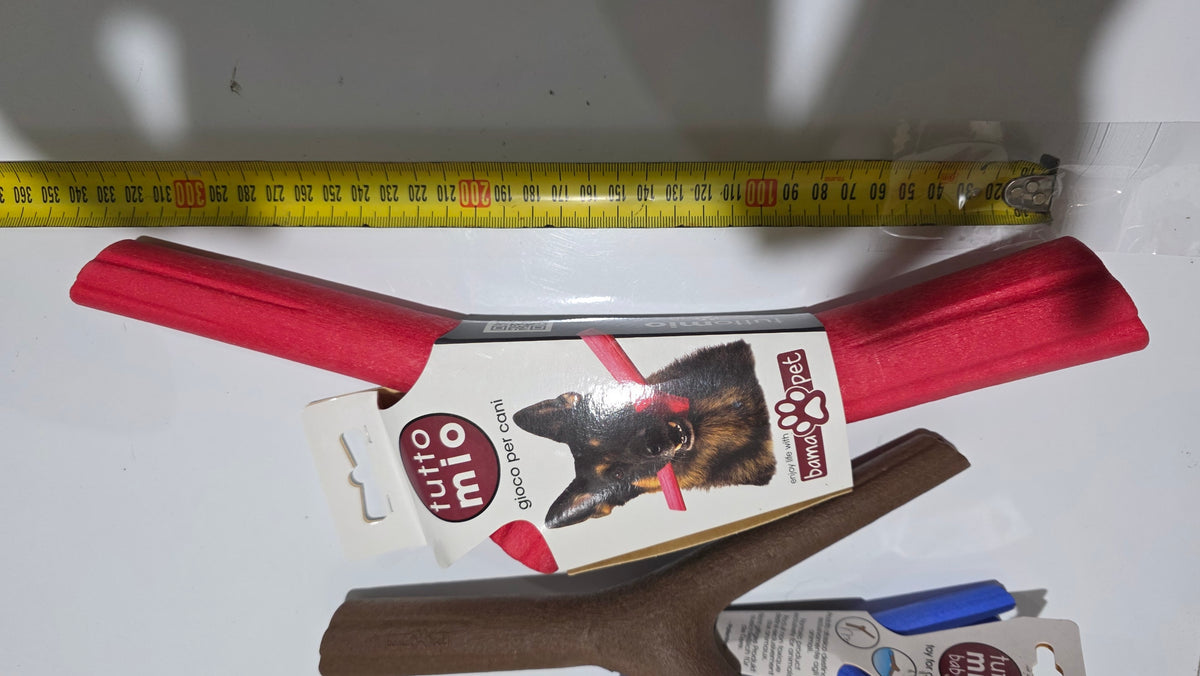 A dog chew toy package labeled 'tutto mio' with red rubber ends and a printed dog photo on the box, photographed on a white surface with a yellow tape measure above and other items partially visible below.