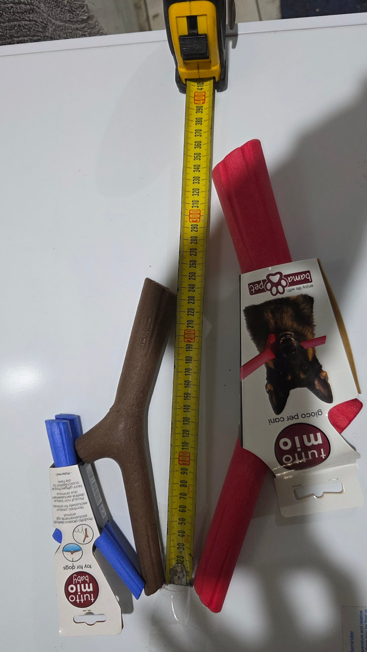 Packaged and unpackaged red, brown, and blue Bama floating stick dog toys arranged on a white surface next to a yellow tape measure demonstrating size differences across small, medium, and large variants.